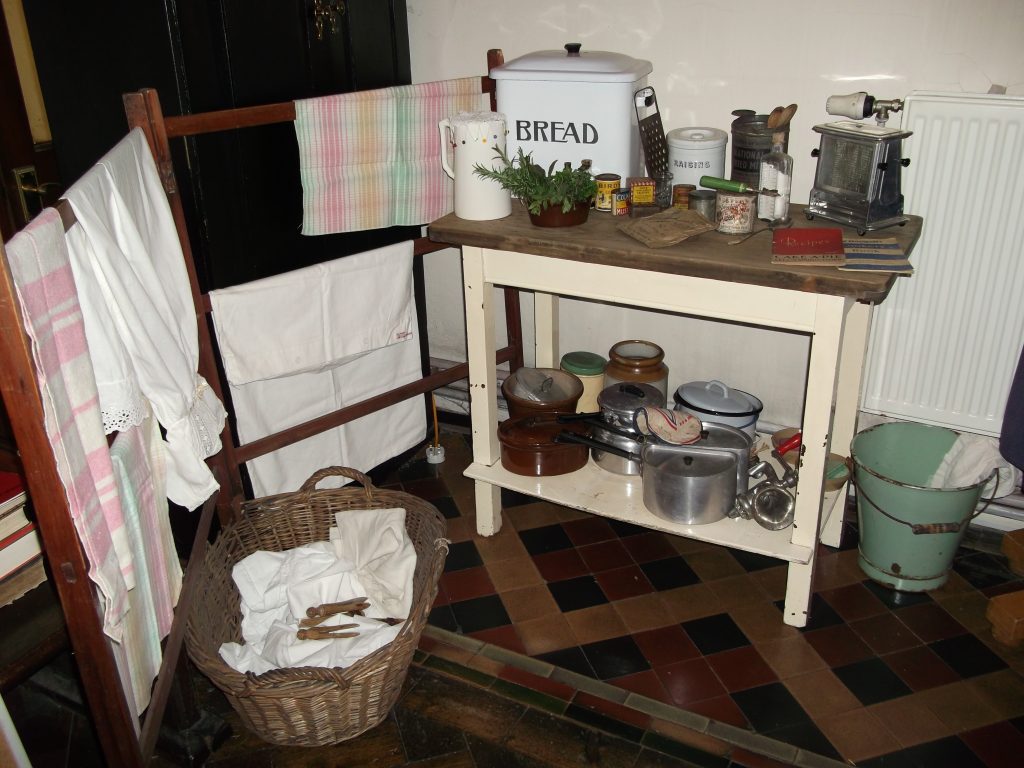 A collection of vintage kitchenalia on a wooden table with a clothes maiden at the side full of vintage household fabrics.