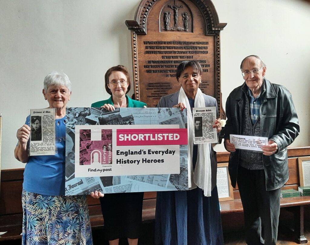 Four church members hold a banner from Heritage Open Days stating 'Shortlisted England's Everyday History Heroes' They are standing in front of a dark wood War Memorial in church